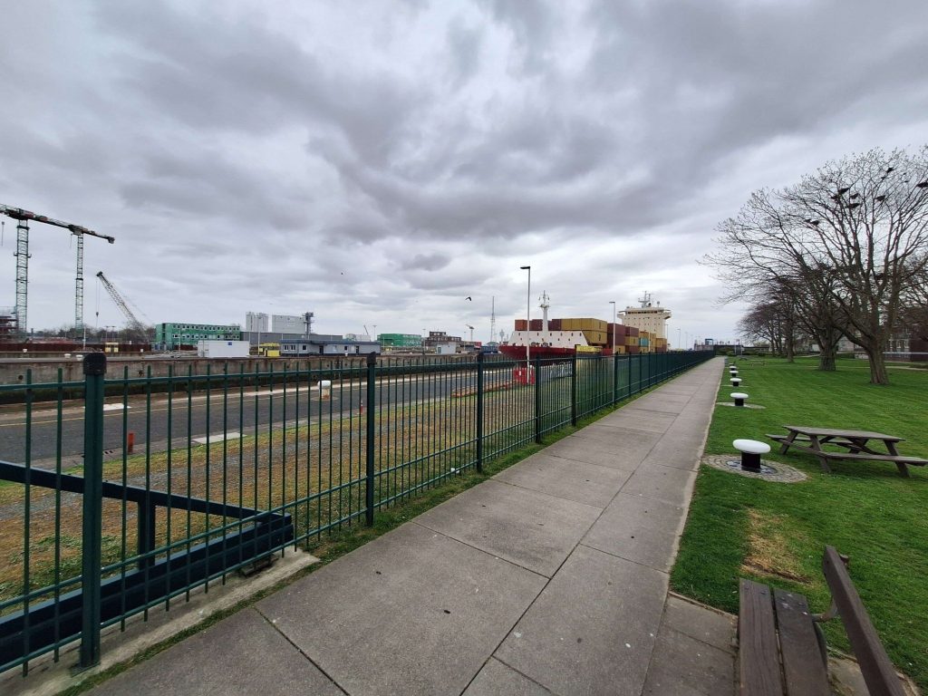 Kanalpromenade in Brunsbüttel mit Containerschiff, Zaun und grüner Wiese – maritime Hafenstimmung auf panikontour.de