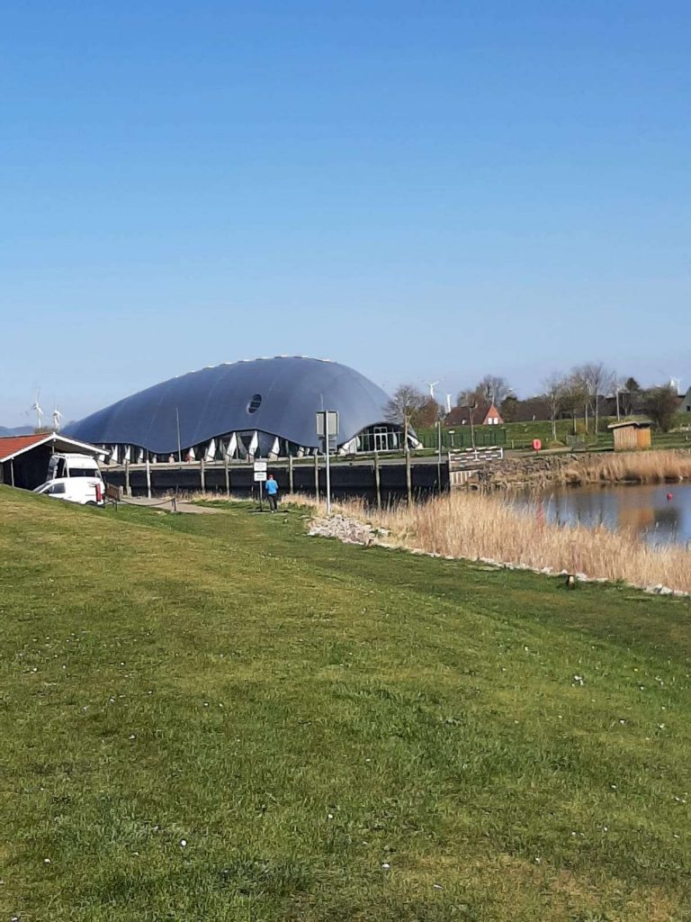 Runde Veranstaltungshalle bei Brunsbüttel mit grünem Deich und blauem Himmel – moderne Architektur in Norddeutschland auf panikontour.de